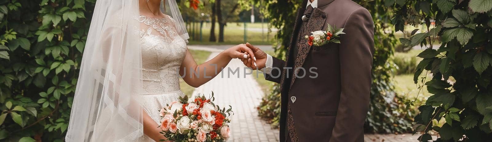 The groom in a brown suit holds the bride's hand with a wedding bouquet by AYDO8