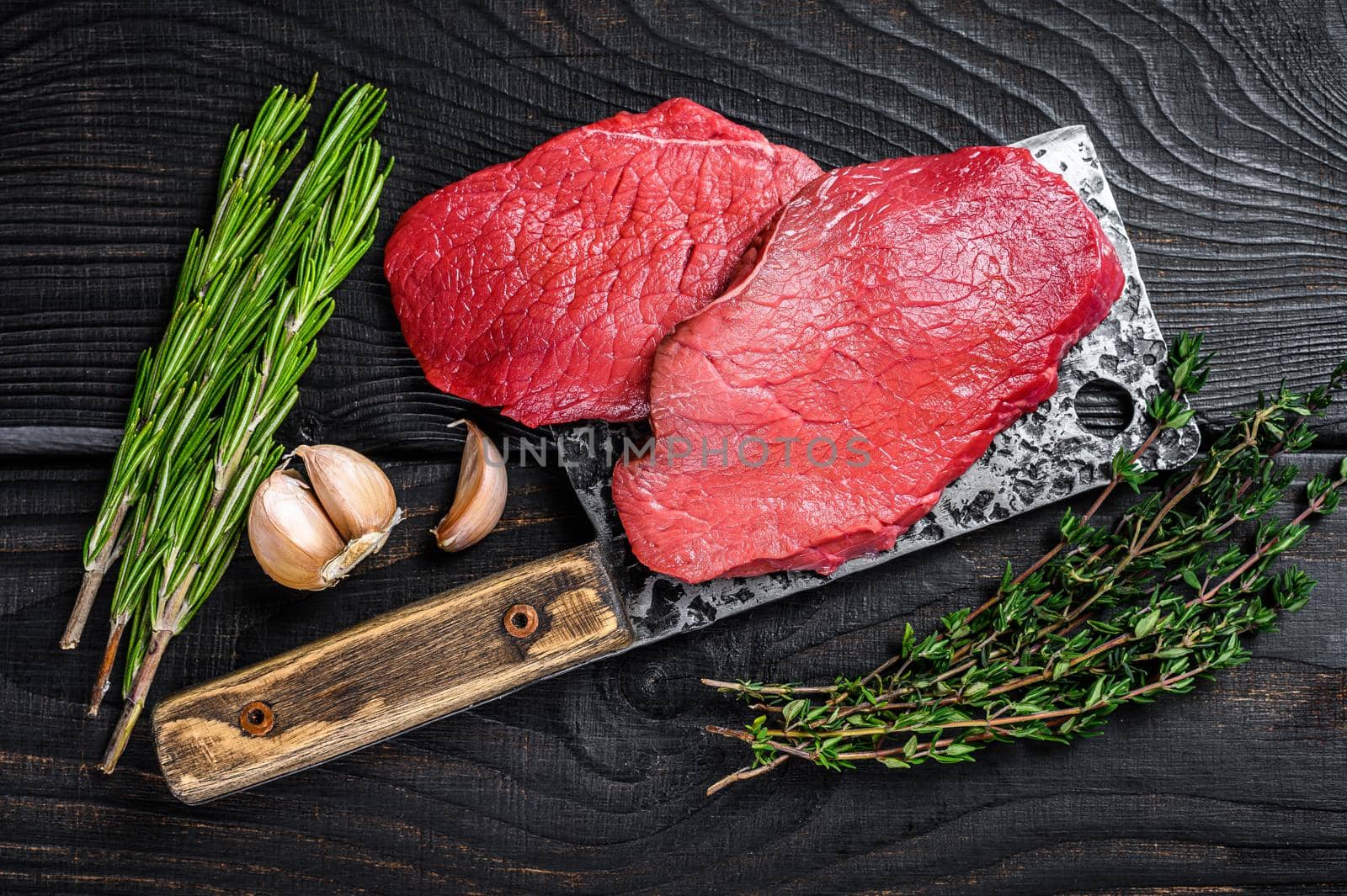 Raw marble beef meat fillet steak on butcher cleaver. Black wooden background. Top view.