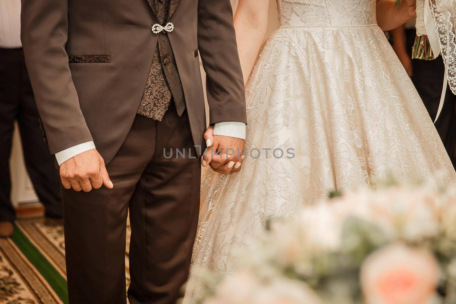 The groom in a brown suit holds the bride's hand tightly.