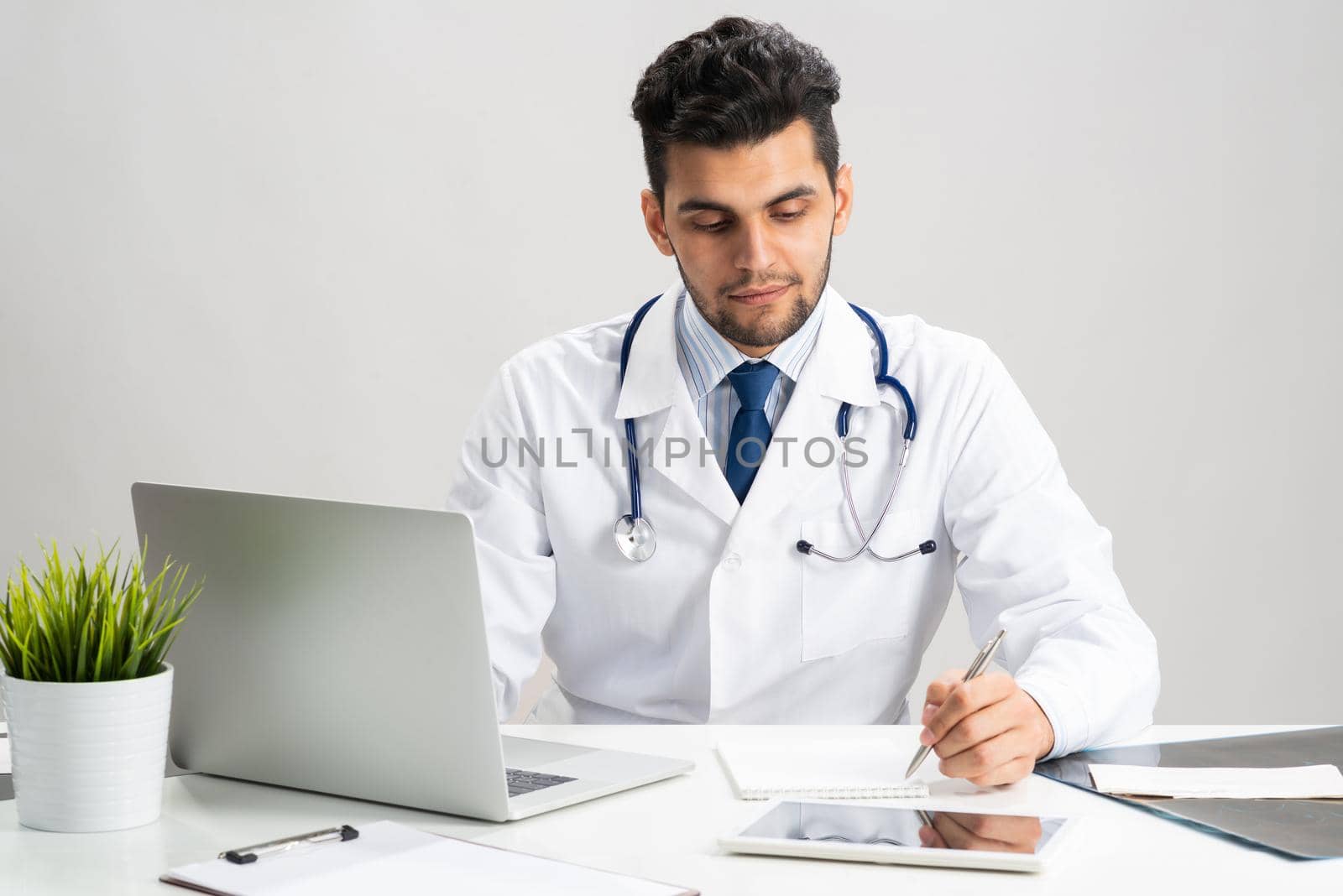 Handsome doctor sitting in office. Young practitioner in white coat with stethoscope at desk with laptop. Professional medical consultation and treatment in modern clinic. Medical healthcare concept