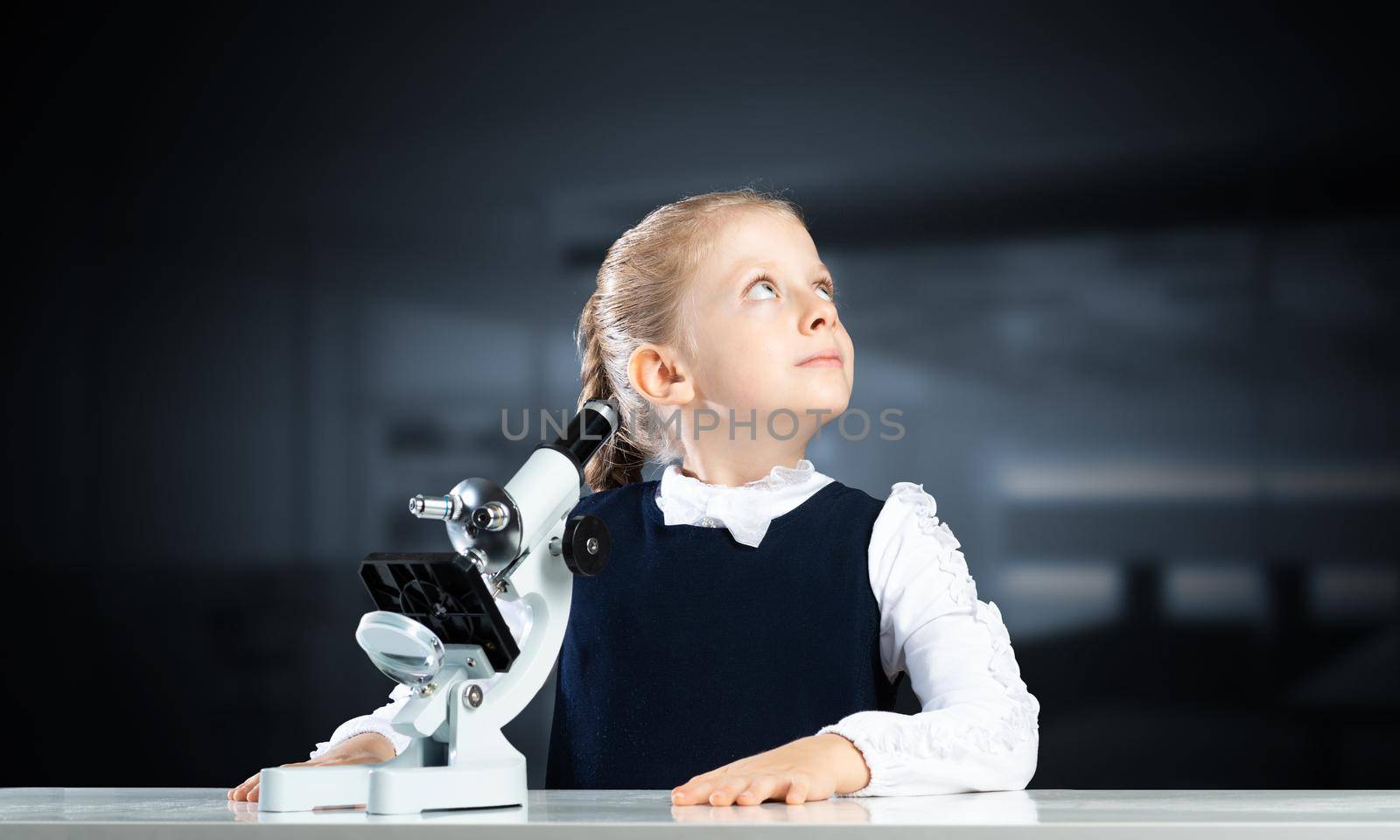 Little girl scientist with microscope on blurred background. Research and discovery concept with copy space. Elementary science class in modern school. Schoolgirl in schoolwear sitting at desk.