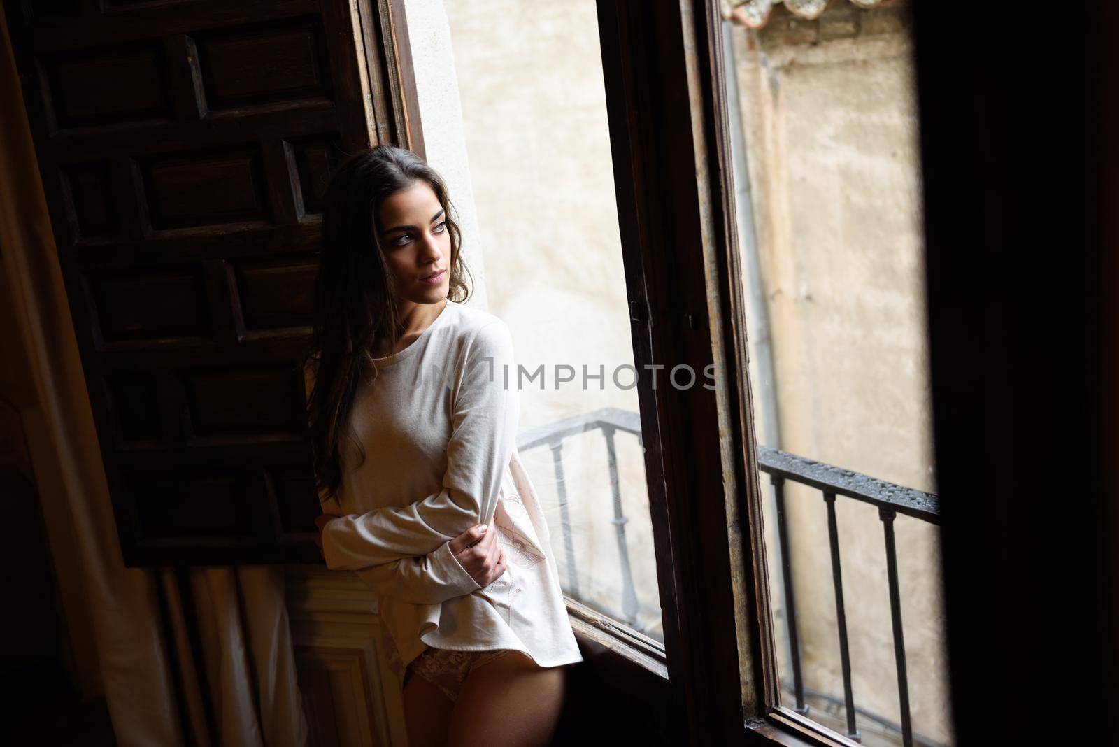 Sexy young woman in pyjama and white panties standing near a window in her bedroom. Brunette girl in lingerie