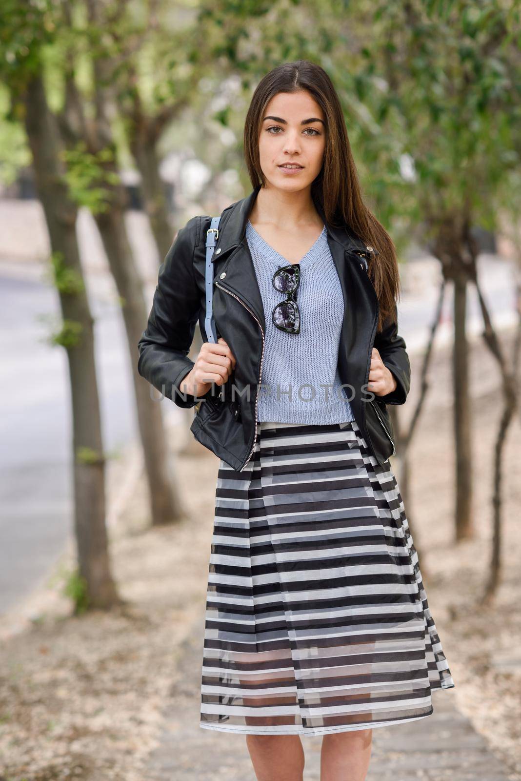 Portrait of young woman smiling in urban background wearing casual clothes. Girl wearing striped skirt, sweater and leather jacket
