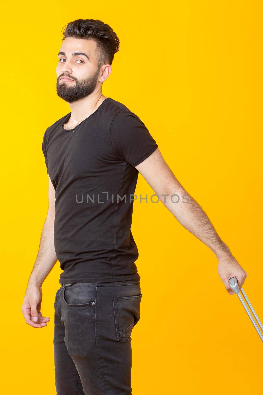 Happy young male hipster with a beard holding a yellow suitcase on a yellow background and rejoicing at a vacation. Travel and tourism concept. Place for advertising