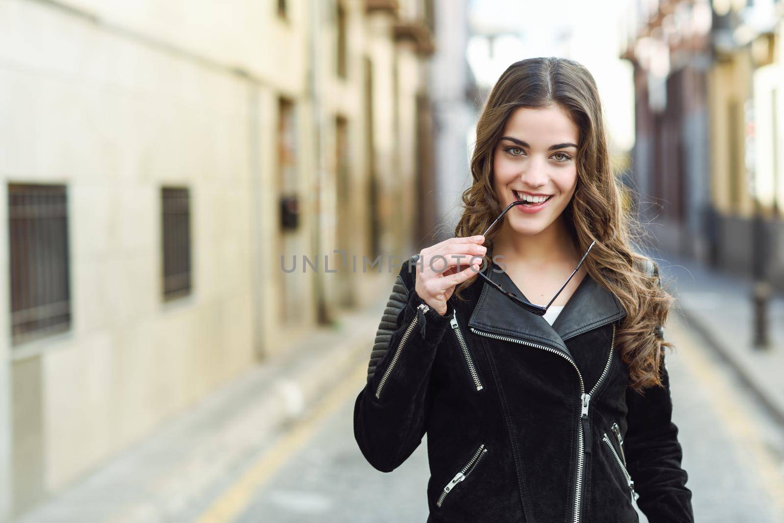 Portrait of young woman smiling in urban background wearing casual clothes with modern sunglasses. Girl with long curly hair