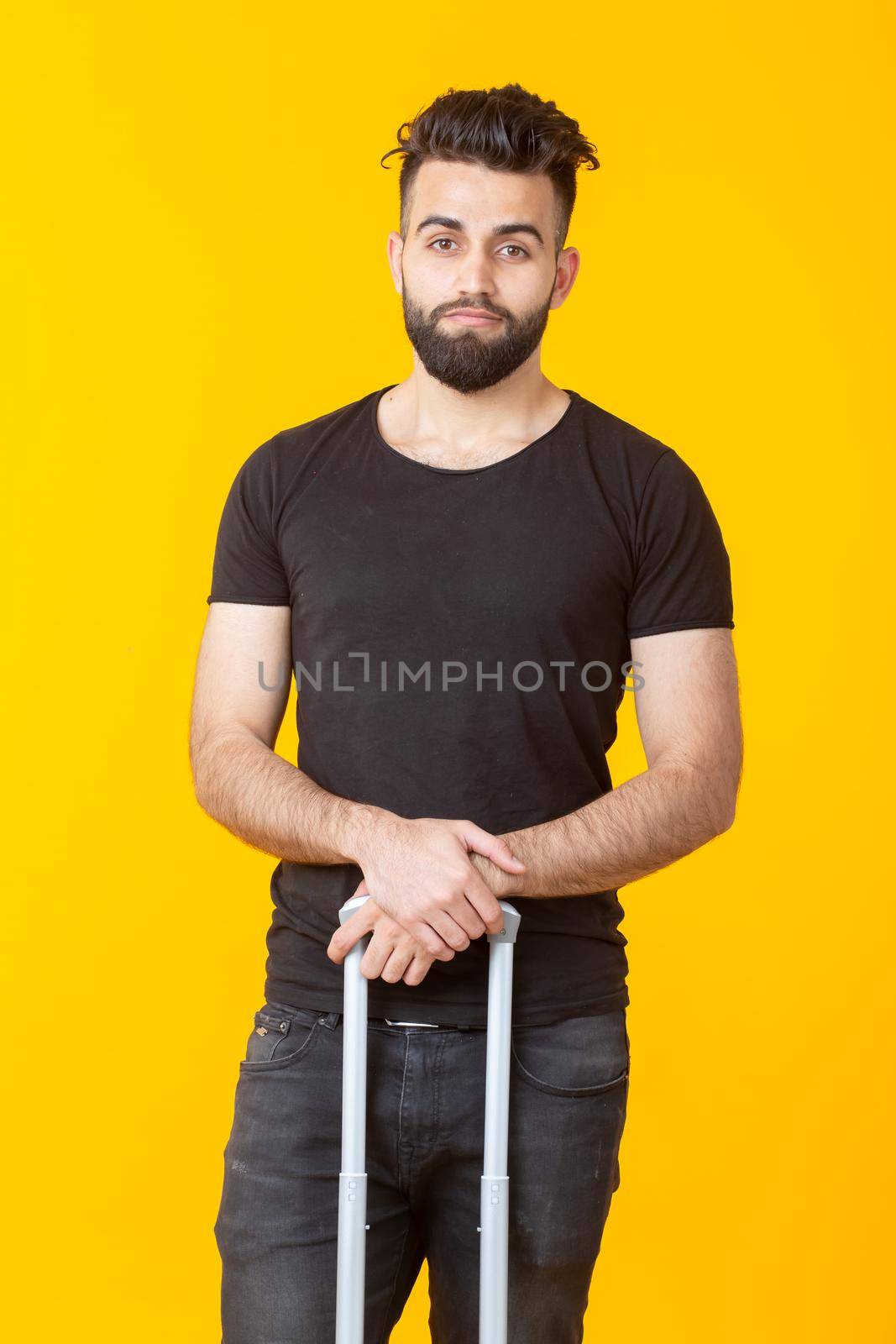 Happy young male hipster with a beard holding a yellow suitcase on a yellow background and rejoicing at a vacation. Travel and tourism concept. Place for advertising