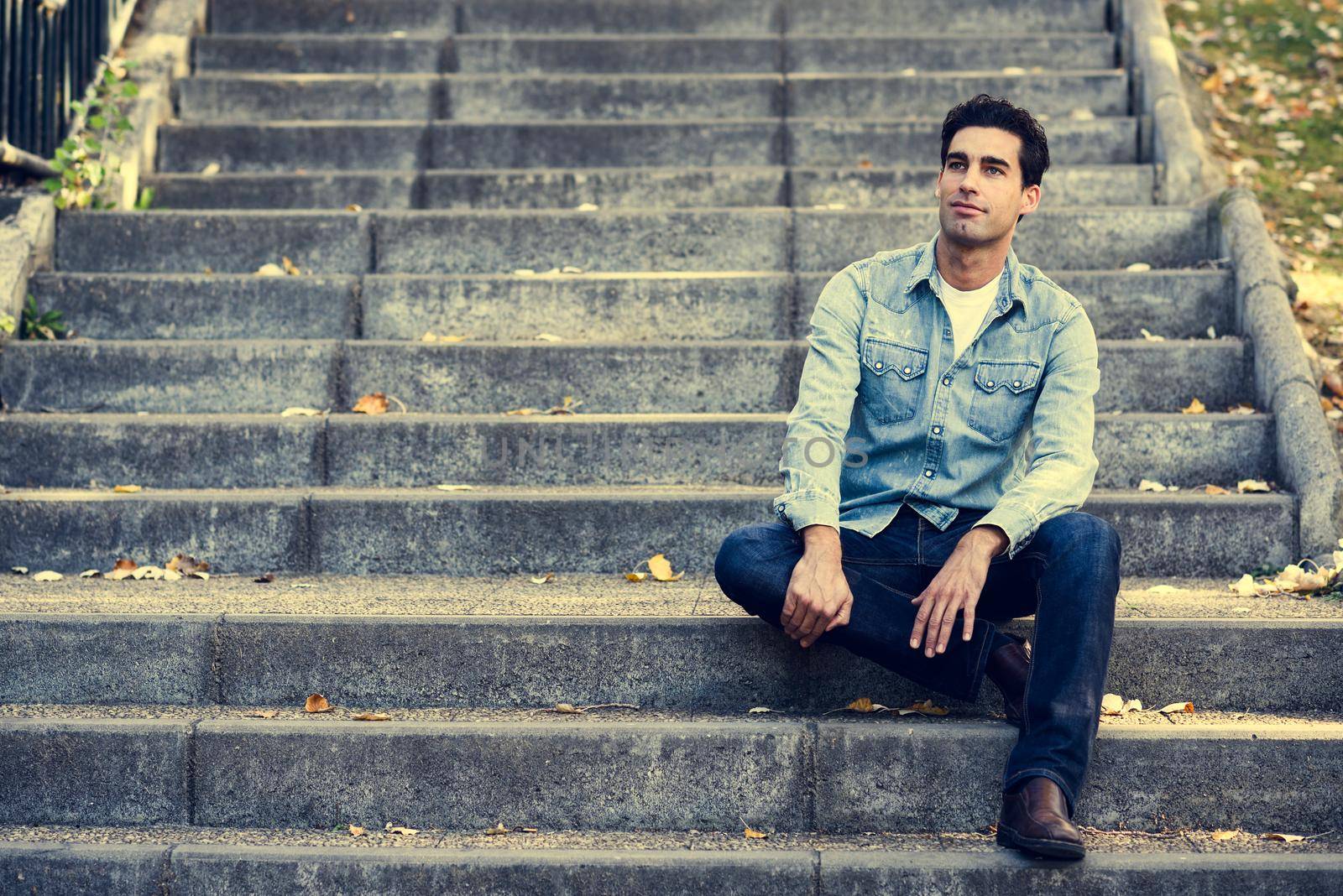 Portrait of a young handsome man, model of fashion, with formal hairstyle sitting in urban stairs, wearing casual clothes. Blue eyes guy in urban background.