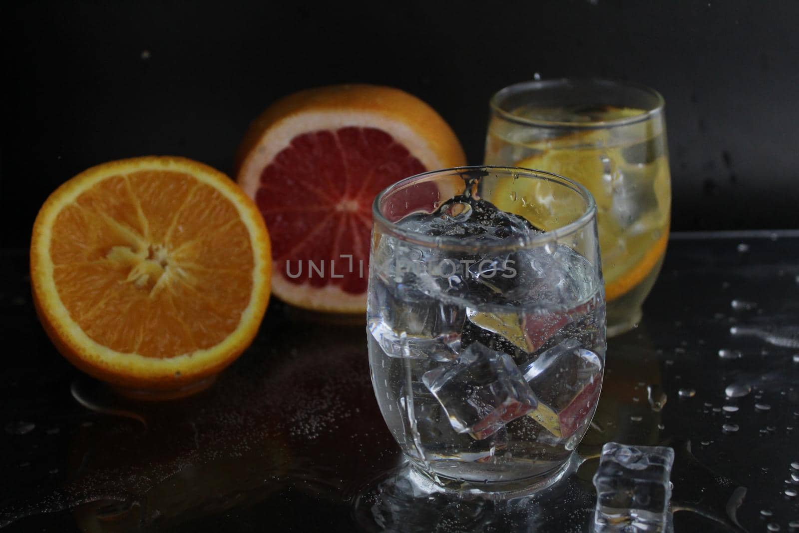 splashes of water over a glass of water and ice. Nearby are fruits orange and grapefruit, ice. On a black background. Refreshing cold ice drink.