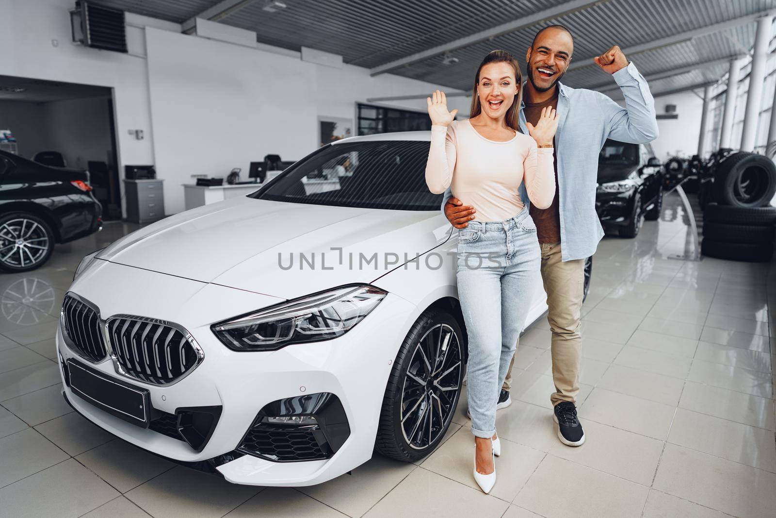 Happy couple of caucasian woman and african american man standing near their new luxury car inside car salon