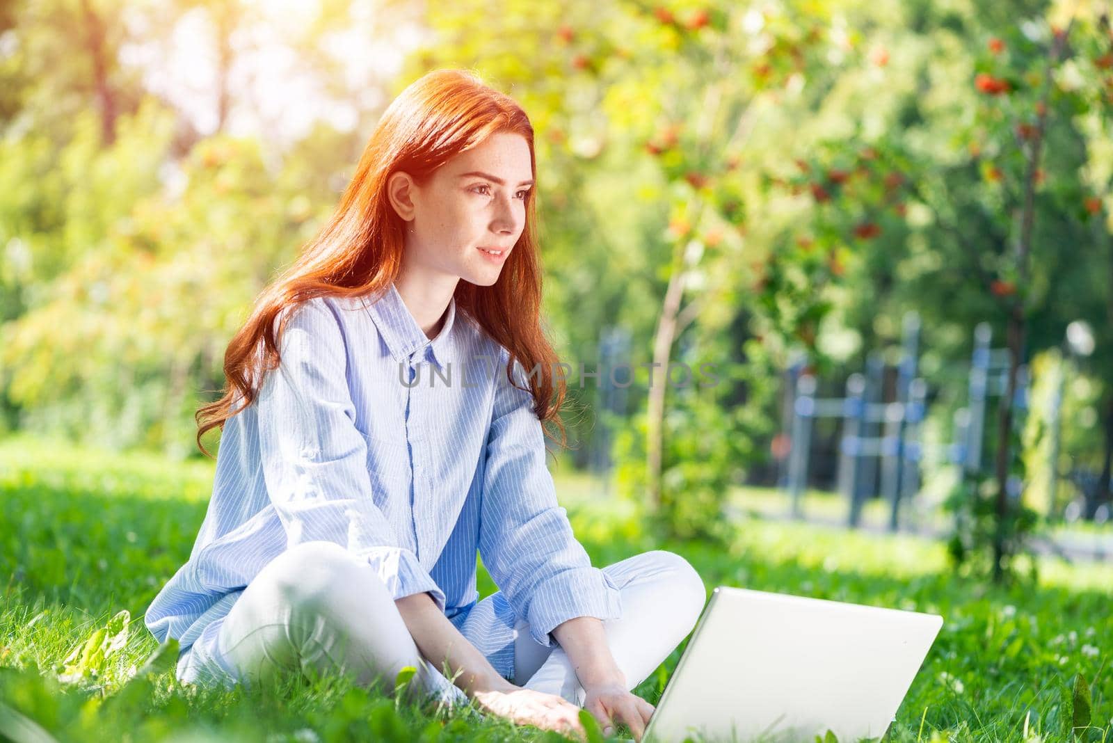 Young redhead woman using laptop computer on green grass in park. Beautiful girl in casual wear working with computer outdoors at sunny summer day. Freelance, blogging or remote work concept.