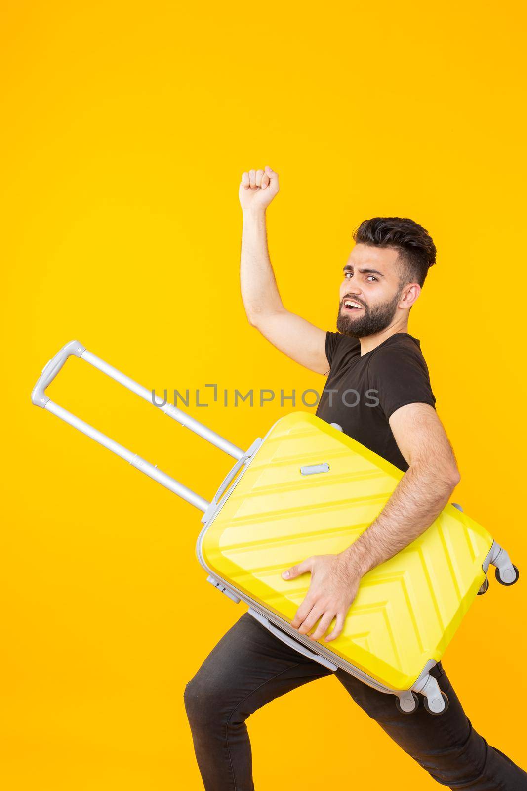 Cute pretty young man with a beard holding a yellow suitcase in his hands on a yellow background. Concept of travel and vacations. Advertising space