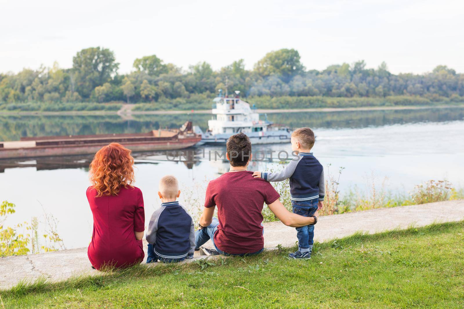 Parenthood, childhood and nature concept - Family sitting on the green ground and looking at small boat by Satura86