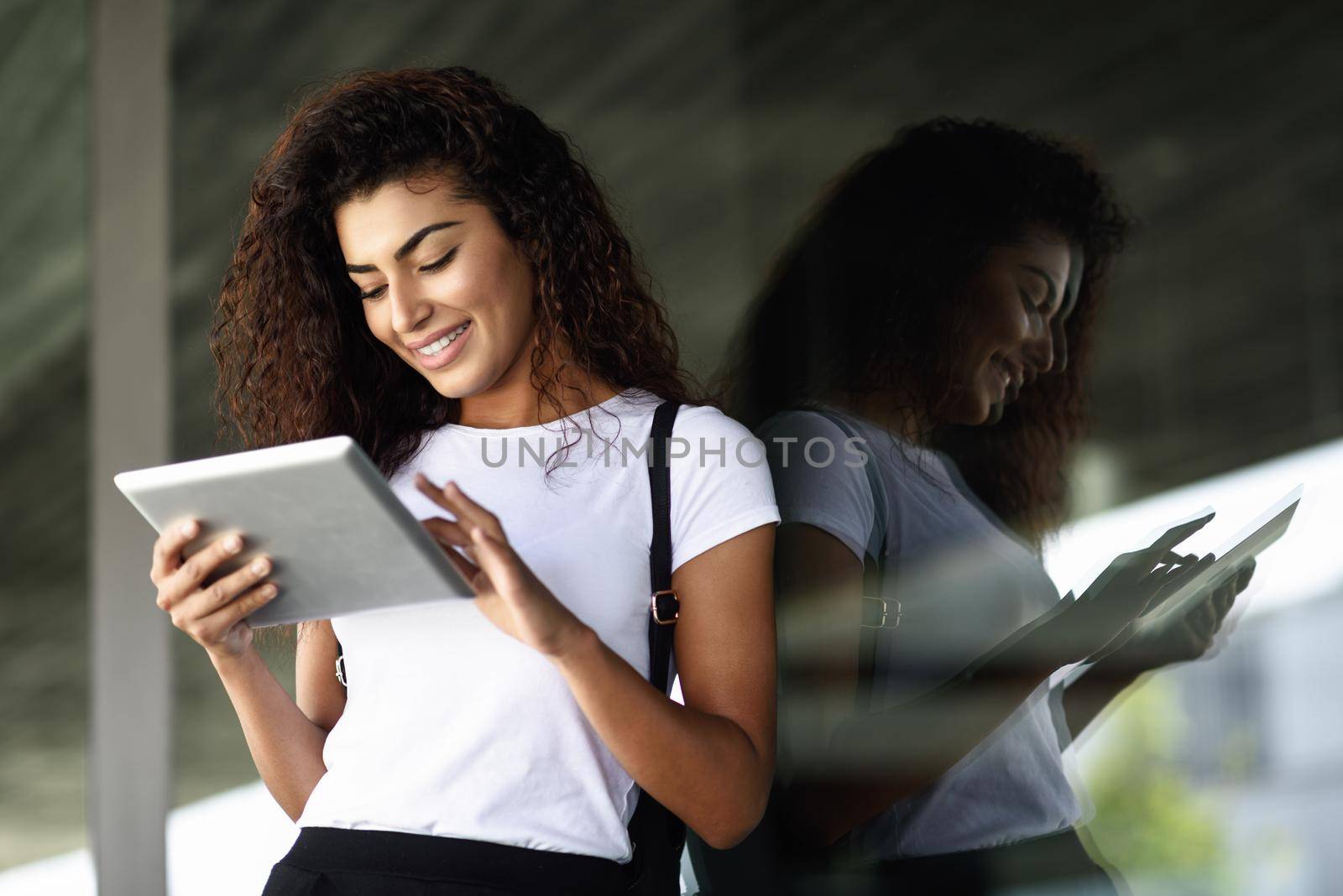 Young North African woman with looking at her digital tablet outdoors. Arab girl using a device reflected in window glass.
