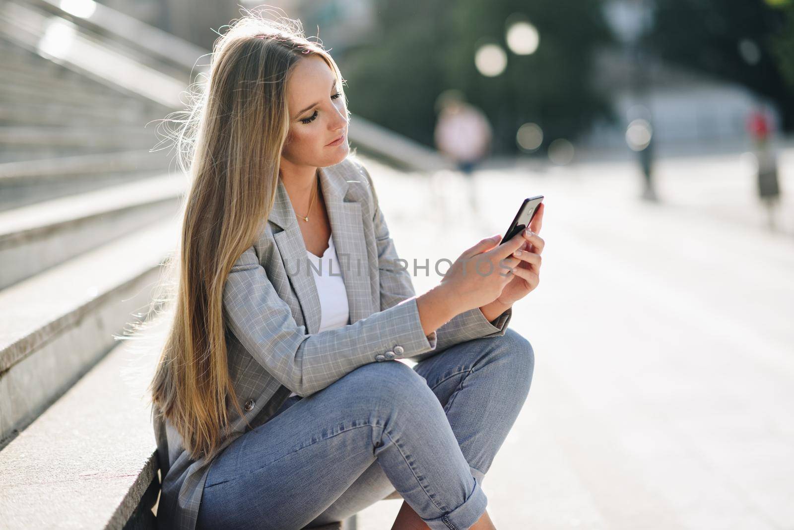 Beautiful young caucasian woman looking at her smartphone in urban background. Blond girl wearing casual clothes sitting on stairs.