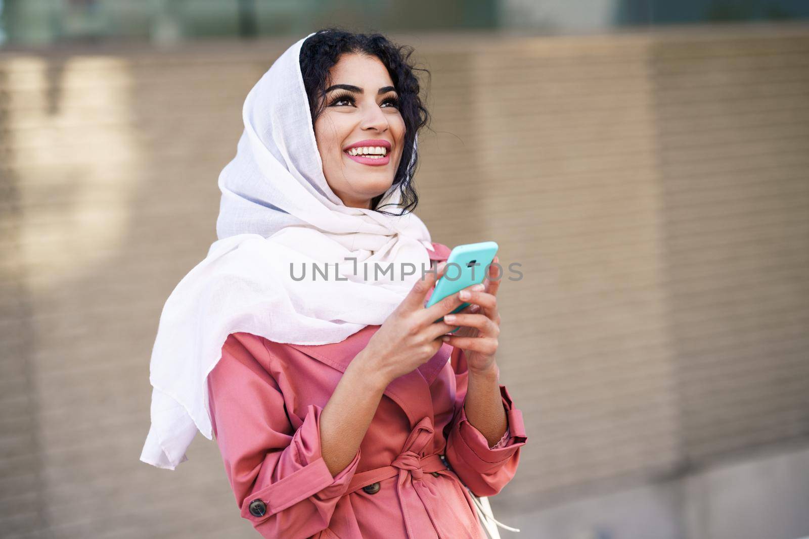 Young Muslim woman wearing hijab headscarf texting message with her smartphone in urban background.