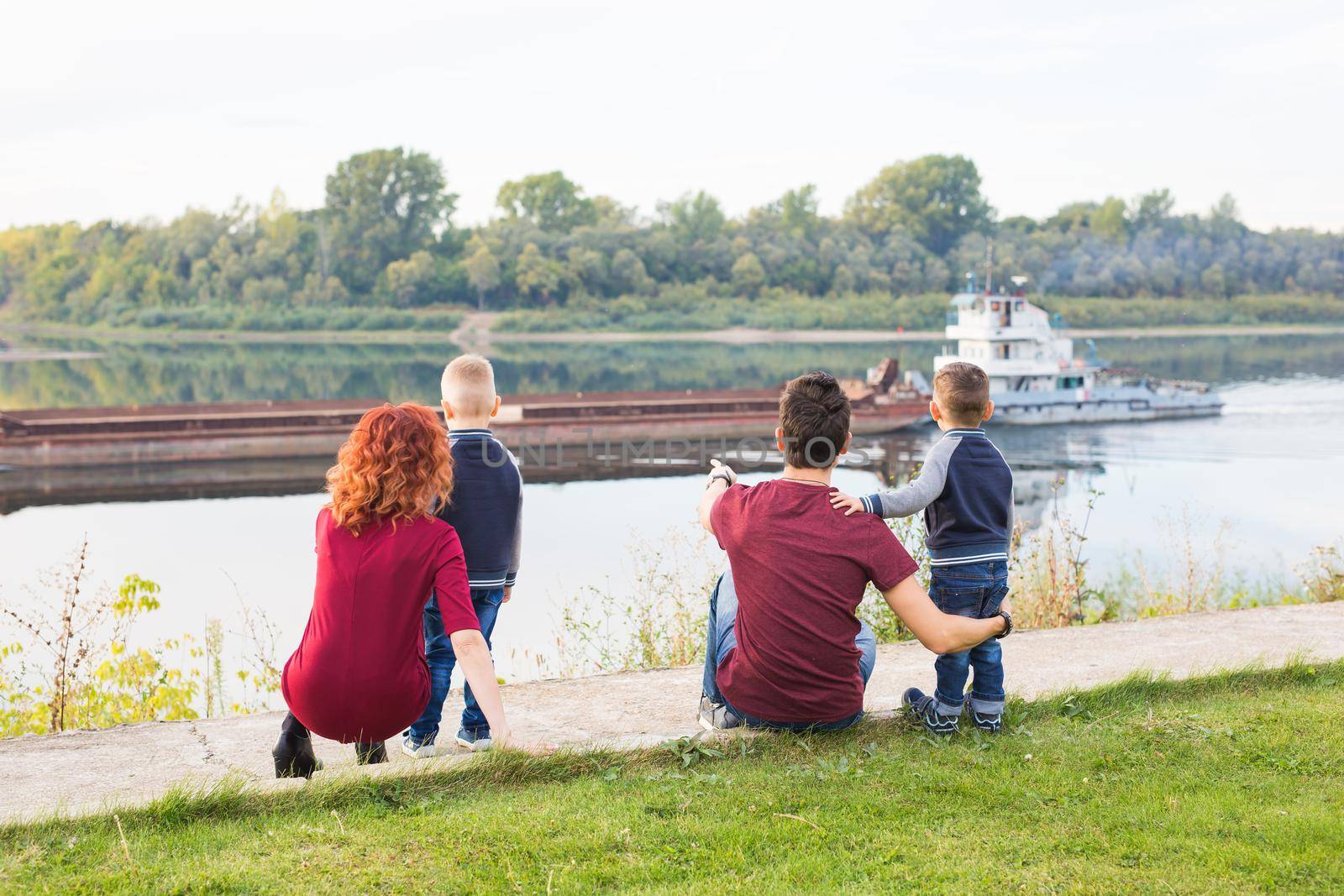 Parenthood, childhood and nature concept - Family sitting on the green ground and looking at small boat by Satura86