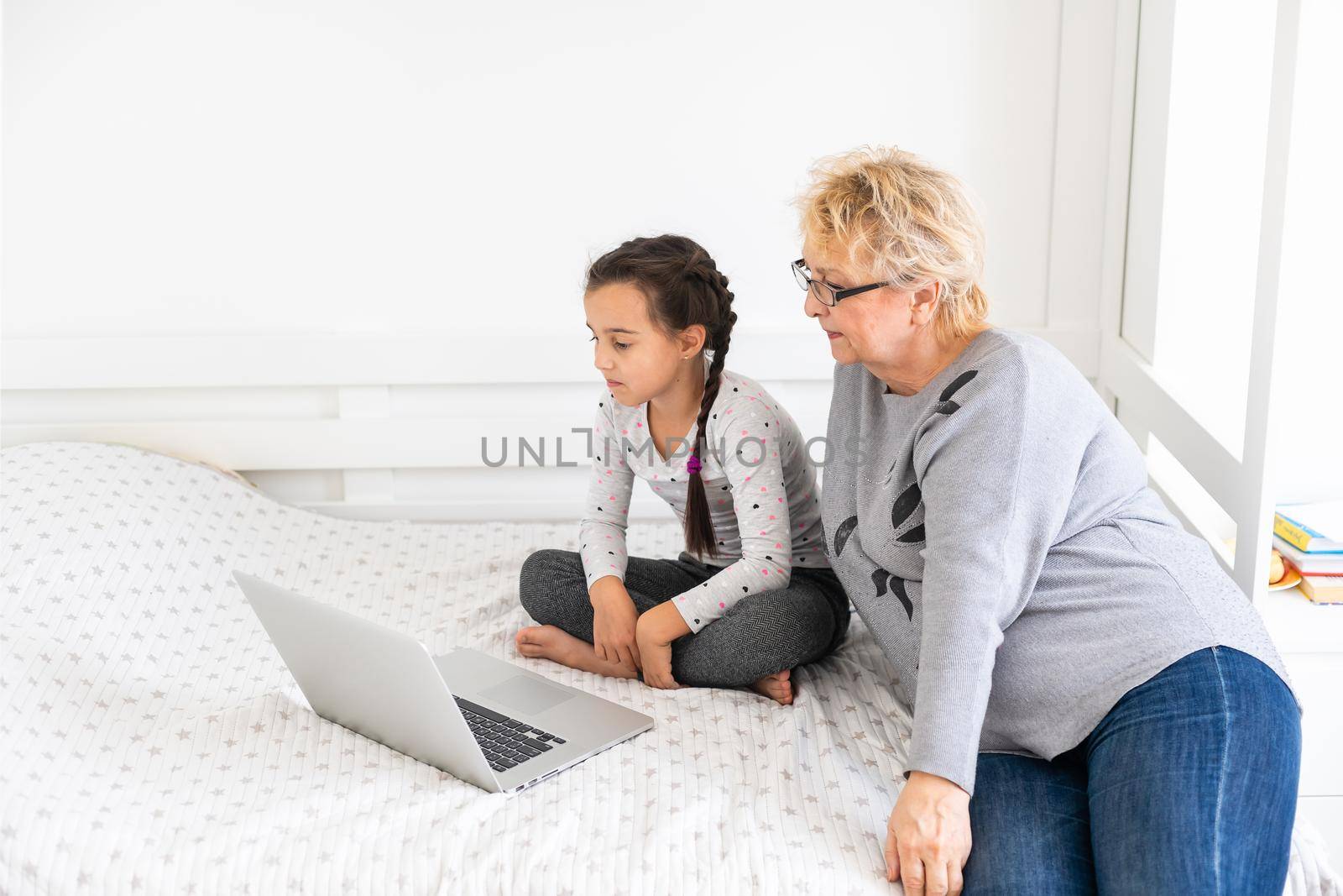 Mature grandmother helping child with homework at home. Satisfied old grandma helping her granddaughter studying in living room. Little girl writing on notebook with senior teacher sitting next to her by Andelov13