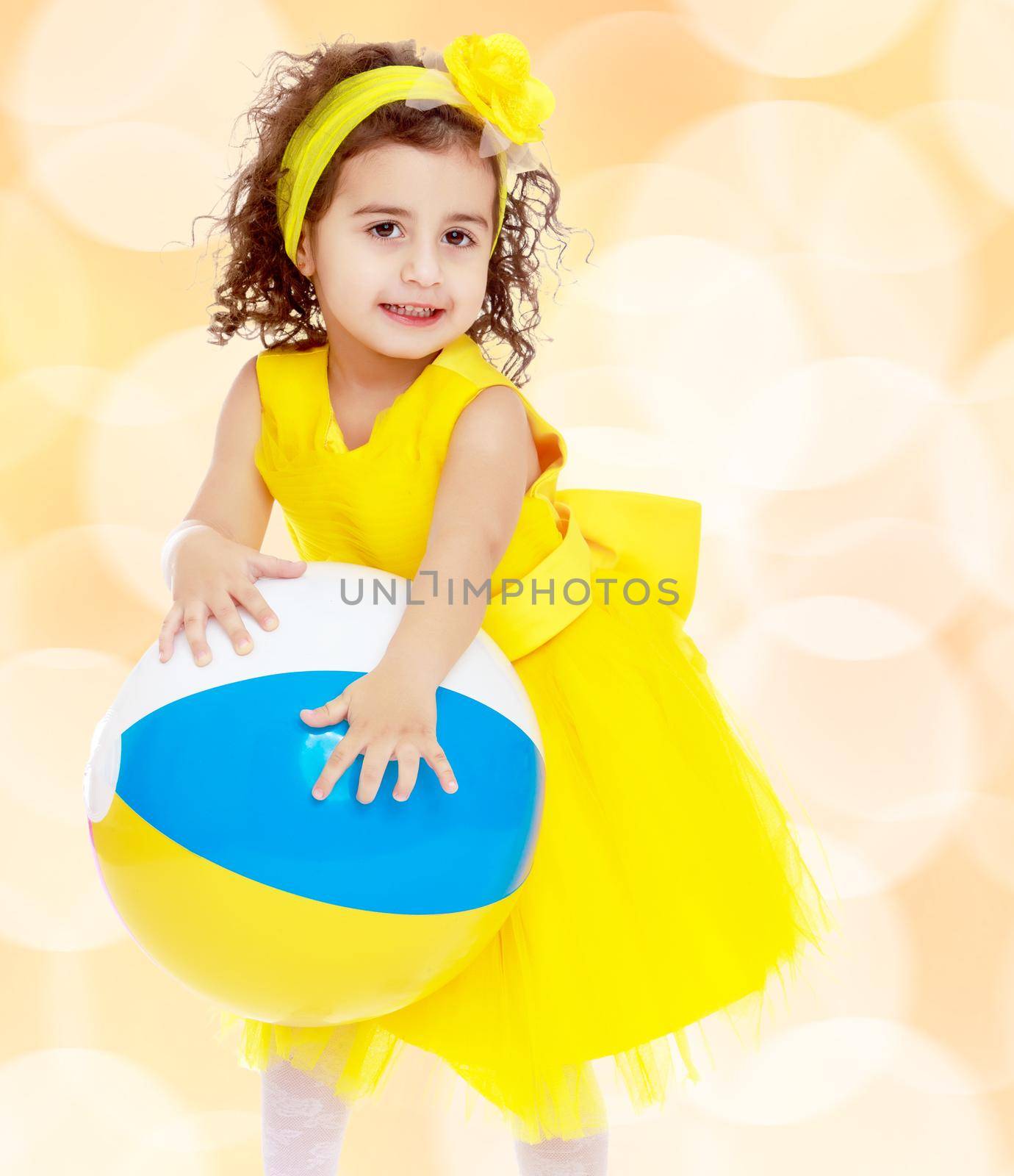 Close-up. Funny curly baby girl in a bright yellow dress and bow on her head playing with a ball.Winter brown abstract background with white snowflakes.
