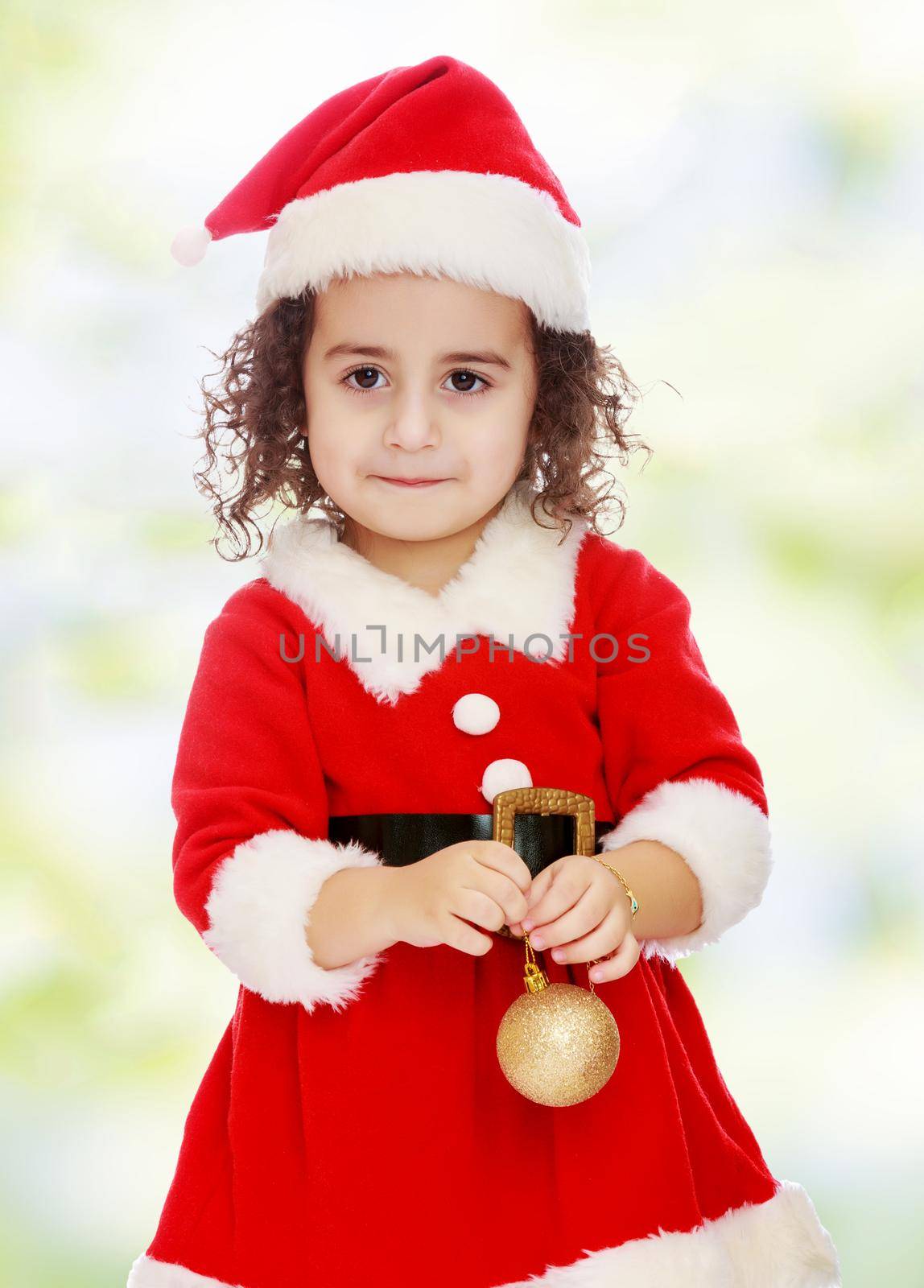 Very cute ,with curly hair little girl in a suit and cap of Santa Claus , is holding a Christmas toy. Close-up.white-green blurred abstract background with snowflakes.