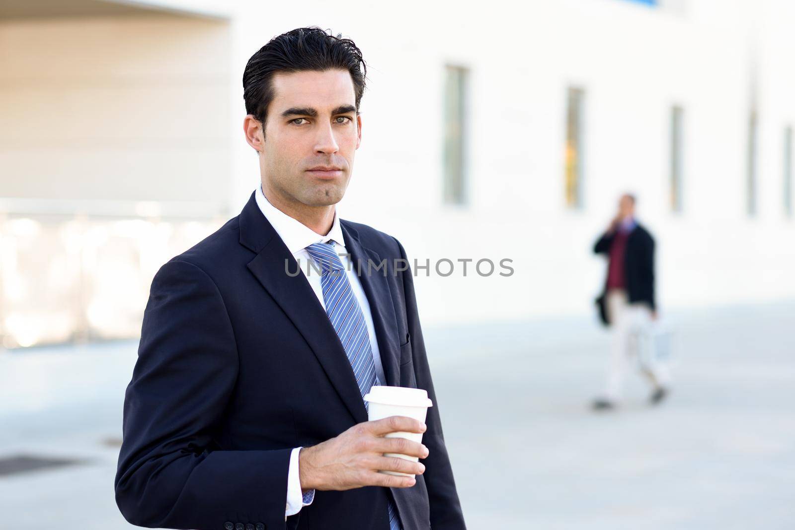 Businessman with formal clothes drinking coffee to go with a take away cup. Man wearing blue suit and tie in urban background.