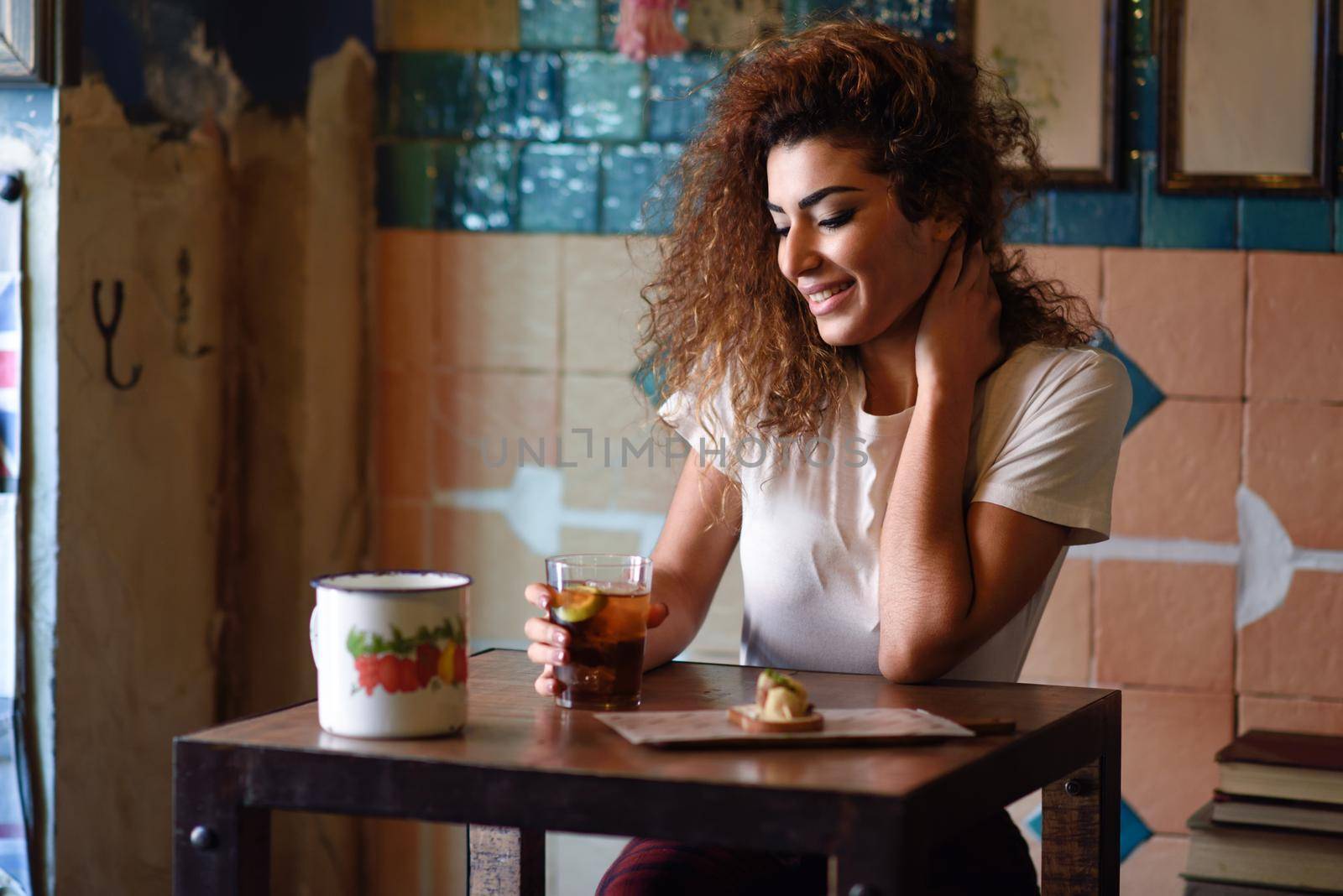 Young arabic smiling woman with black curly hairstyle sitting in a beautiful bar with vintage decoration. Arab girl in casual clothes drinking a soda.