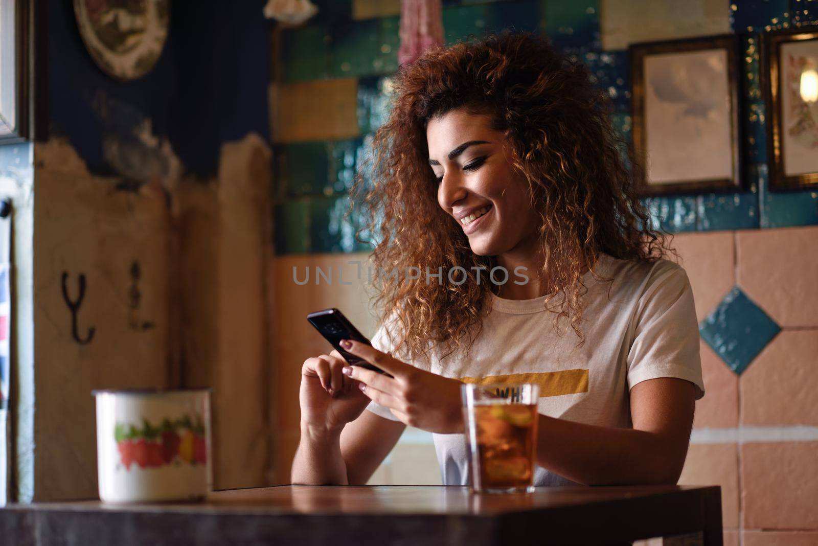 Young arabic woman with black curly hairstyle sitting in a beautiful bar with vintage decoration. Arab girl in casual clothes drinking a soda and looking at her smart phone