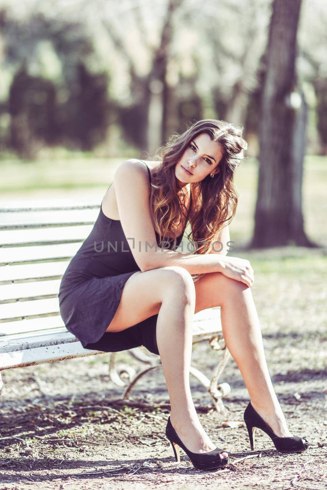 Portrait of a pretty woman sitting in a bench in a urban park wearing black dress and high heels