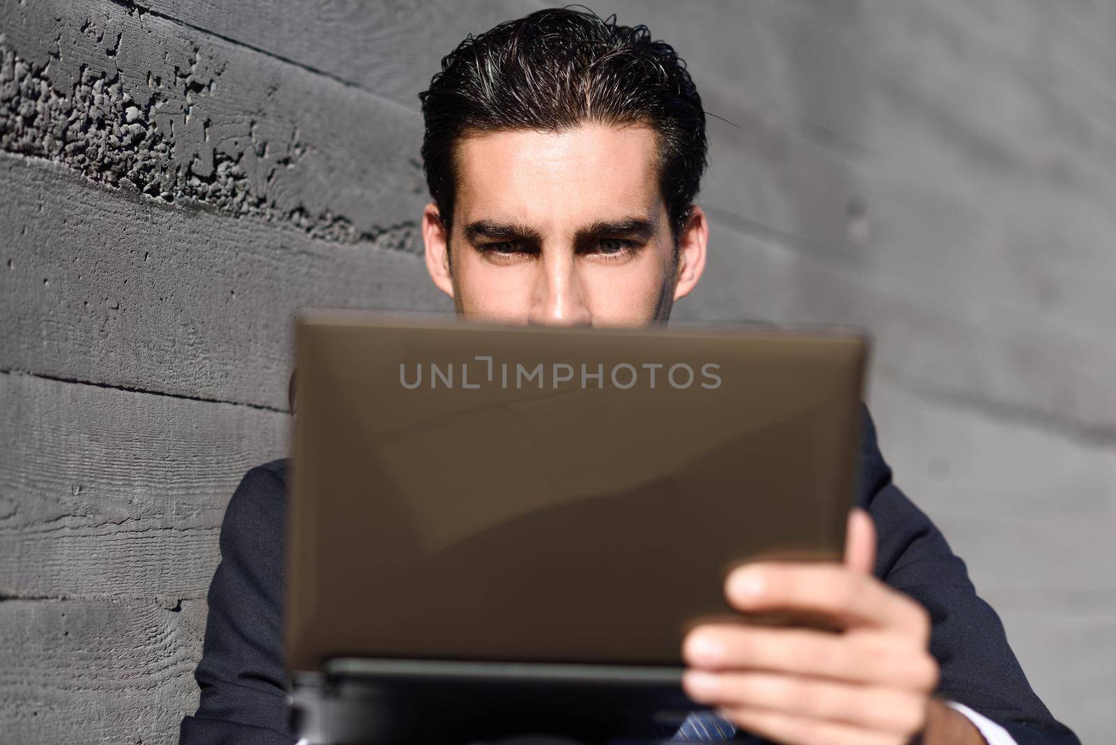 Good looking businessman using a laptop computer sitting in the street. Man wearing blue suit and tie in urban background.