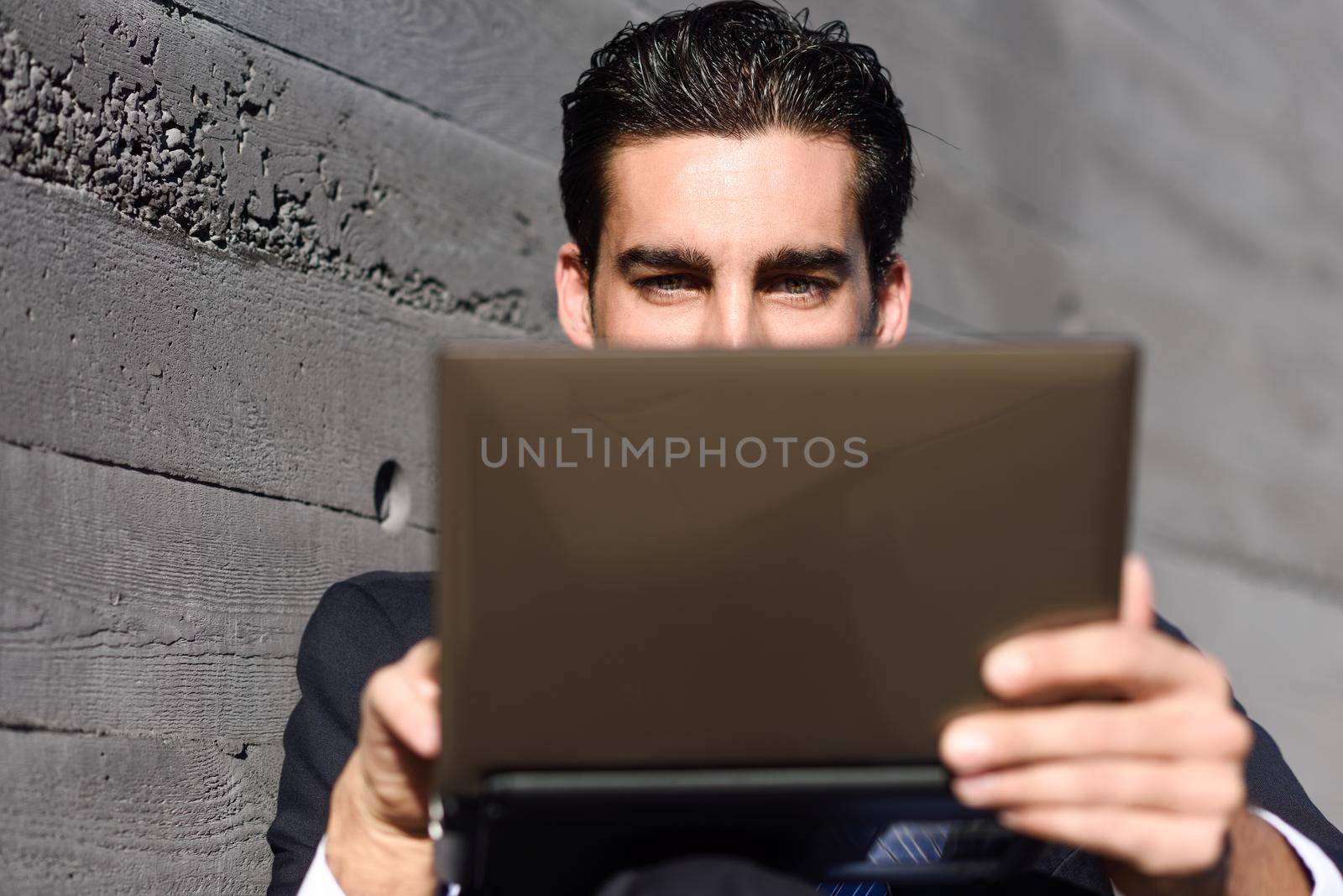 Good looking businessman using a laptop computer sitting in the street. Man wearing blue suit and tie in urban background.