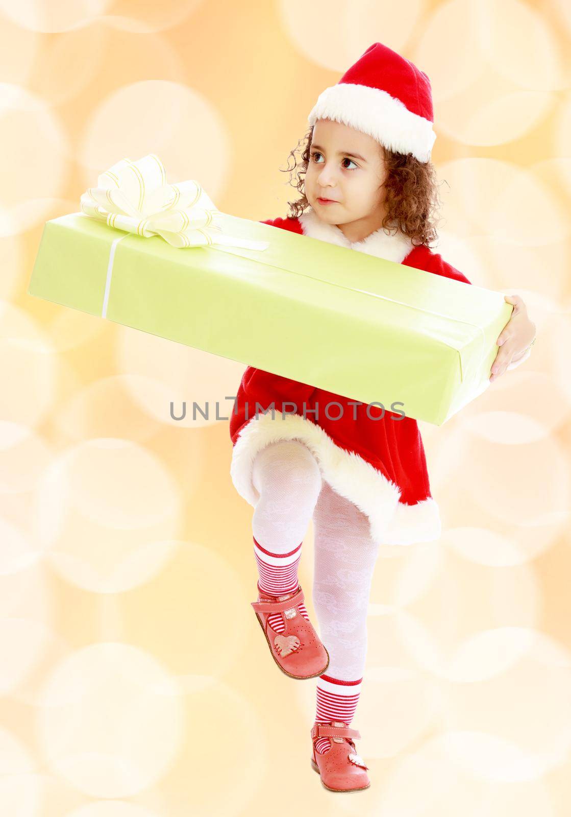 Cute little curly girl in a coat and hat of Santa Claus carries the big green box. A girl holds a box with his knee.Winter brown abstract background with white snowflakes.