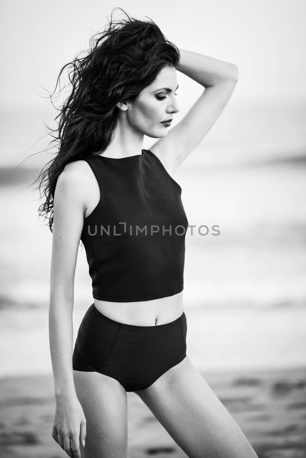 Brunette woman, model of fashion, wearing black top and panties in front of a beach. Young girl with curly hairstyle moving hair.