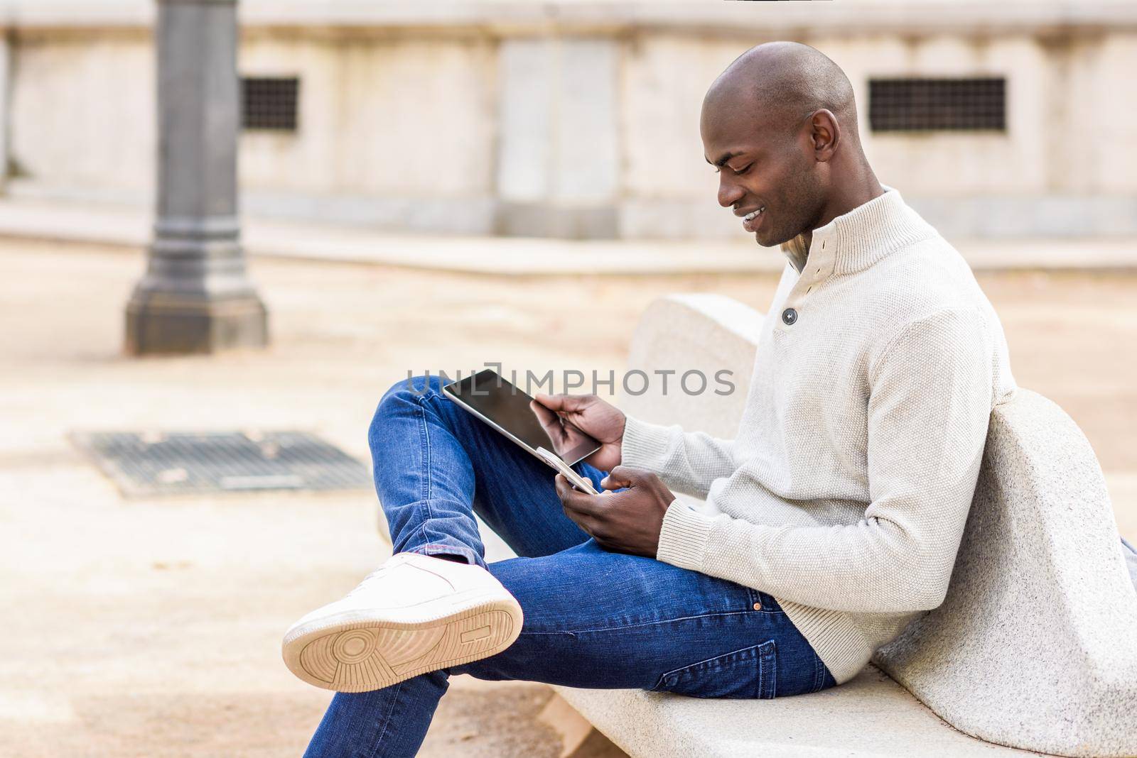 Black young man looking at tablet computer and smartphone in urban background. Young african guy with shaved head wearing casual clothes.