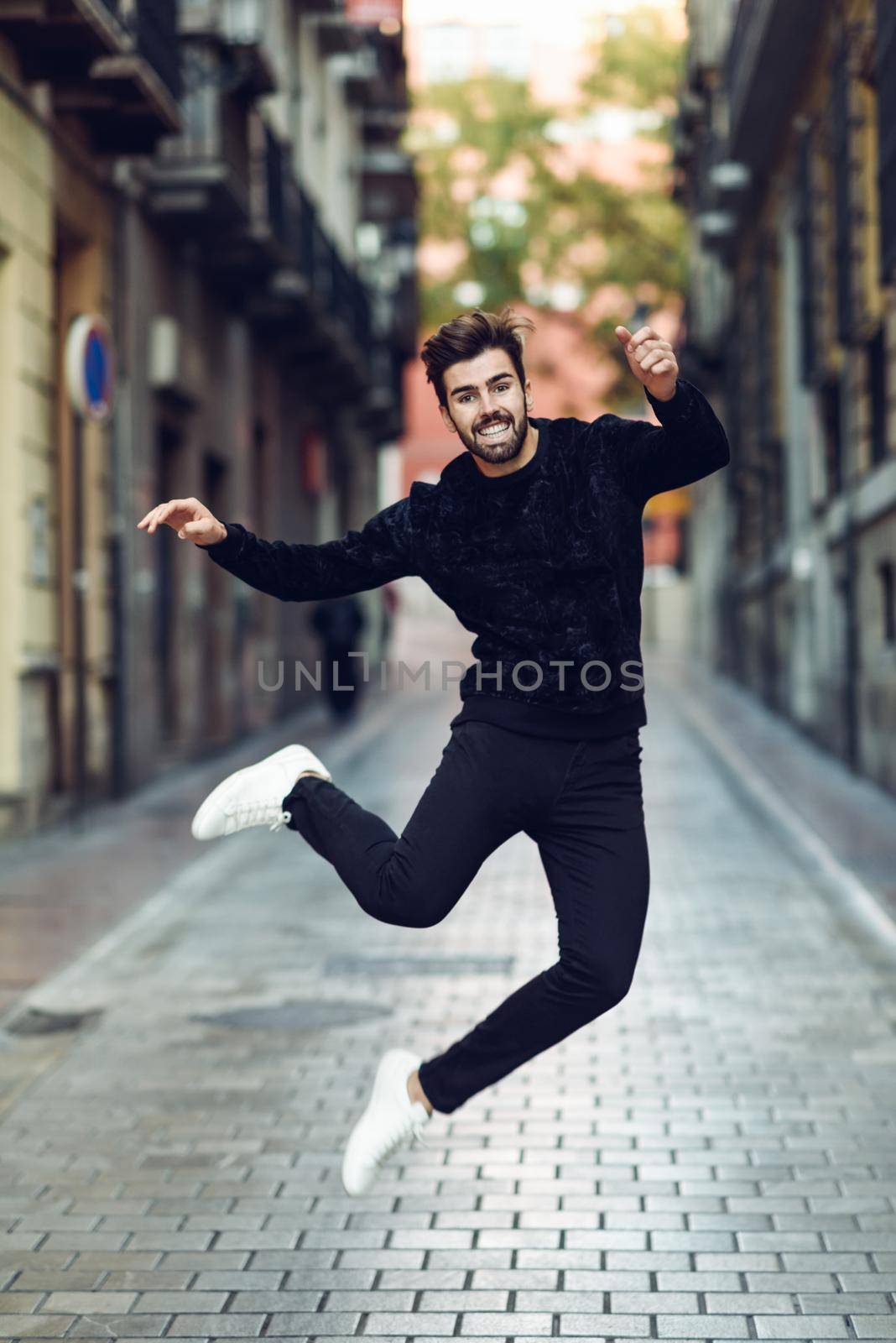 Young bearded man jumping in urban street wearing casual clothes. Happy guy with beard and modern hairstyle.