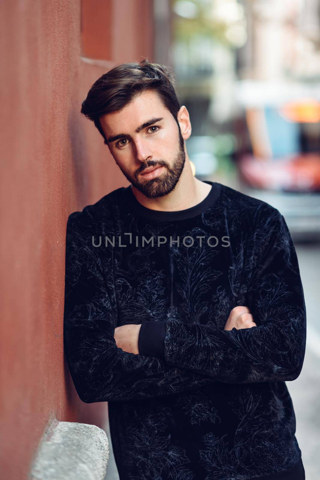 Young bearded man, model of fashion, looking at camera in urban background wearing casual clothes. Guy with beard and modern hairstyle in the street.