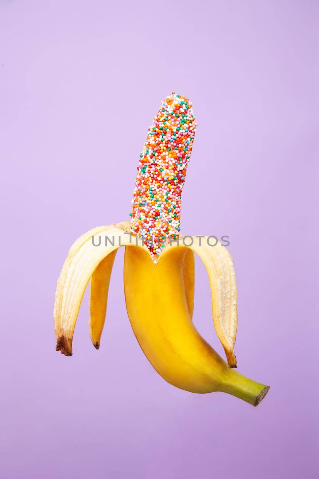 Yummy peeled banana covered with sweet multicolored sprinkles and isolated on purple background