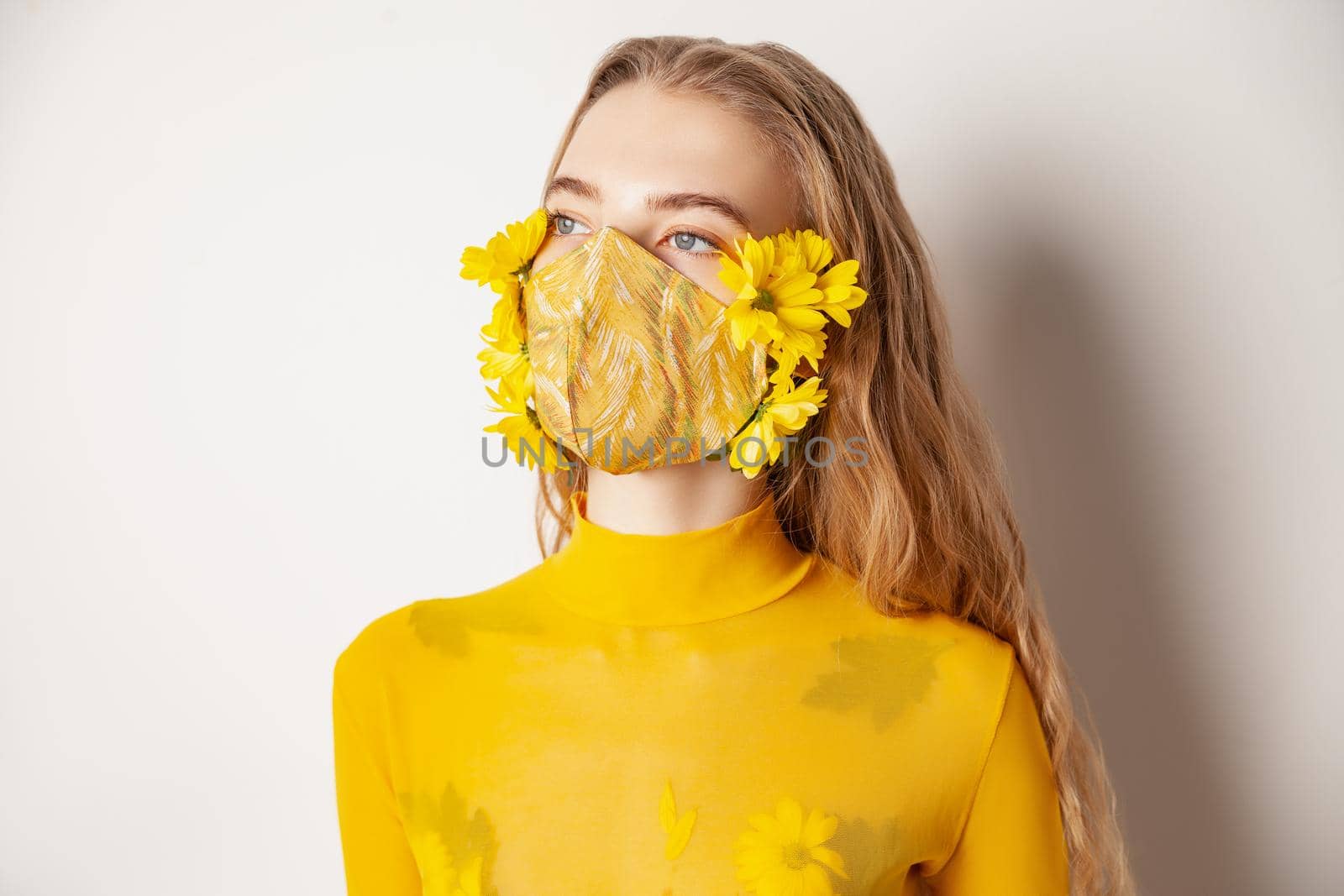 Slim female model in protective mask with fresh yellow flowers standing in transparent outfit on white background in studio
