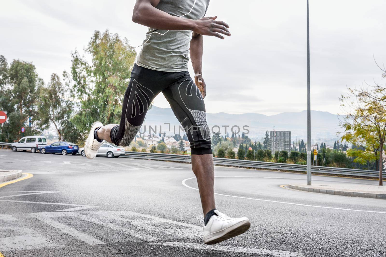Black man running outdoors in urban road listening to music with white headphones. Young male exercising with city scape at the background.