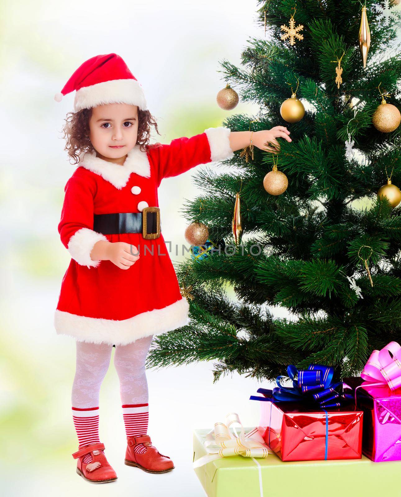Adorable little curly-haired girl, dressed as Santa Claus decorates a Christmas tree toys.white-green blurred abstract background with snowflakes.