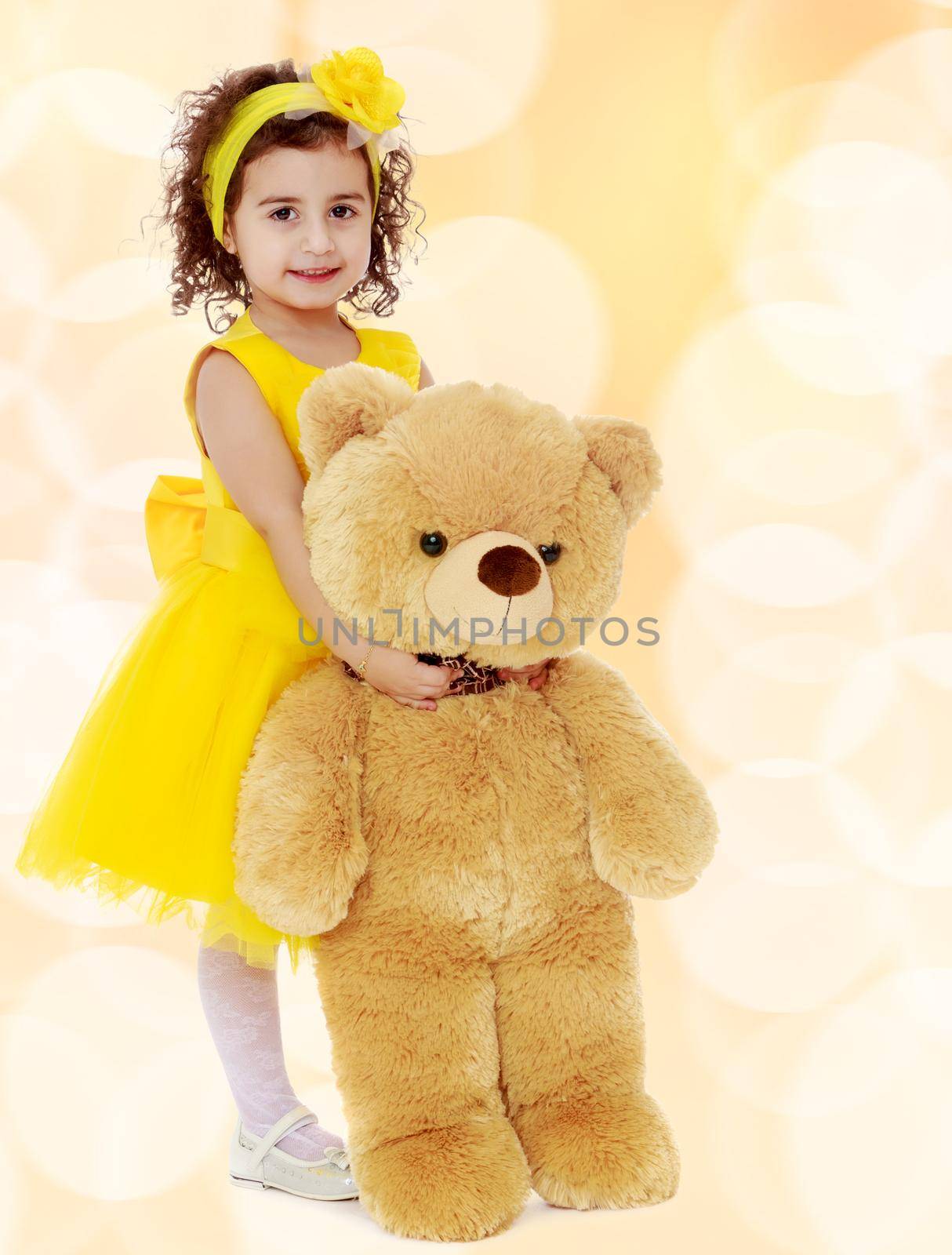 Joyful little girl Princess, dressed in a yellow dress, hugging a big Teddy bear. Standing at full height.Winter brown abstract background with white snowflakes.