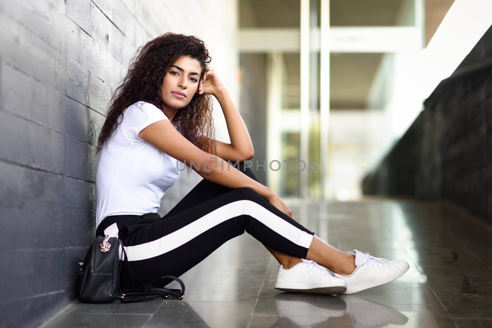 African woman with black curly hairstyle sitting on urban floor. Arab girl in sport clothes in the street.