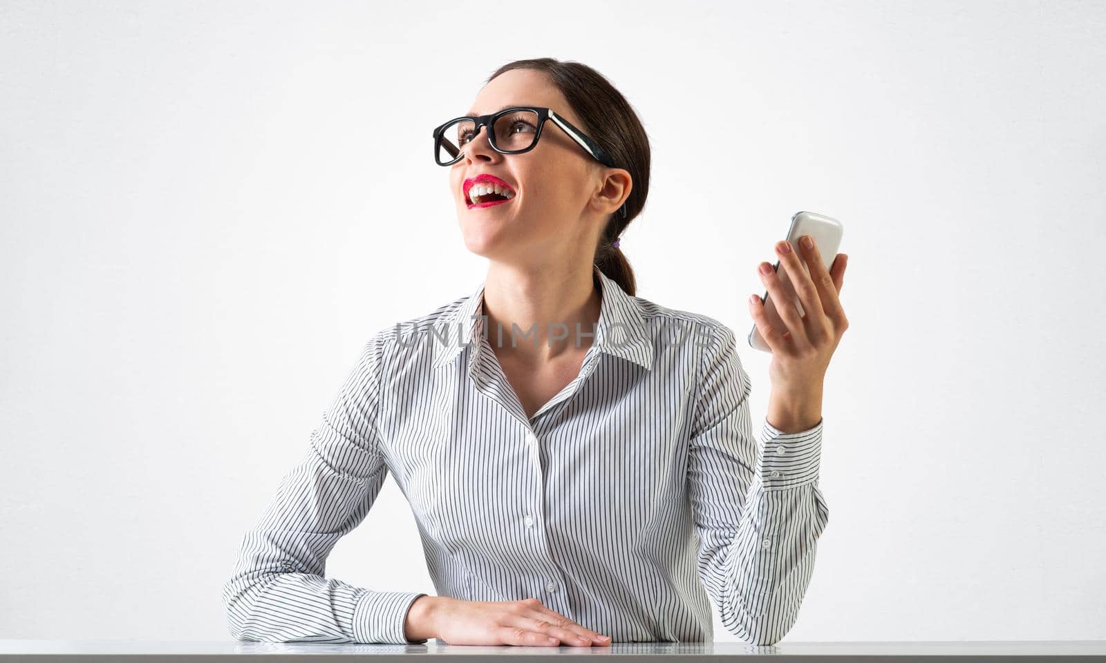 Smiling young woman sits at desk and dreamy looking upward. Portrait of beautiful girl in glasses with bright red lips on white wall background. Human resource and career development concept.