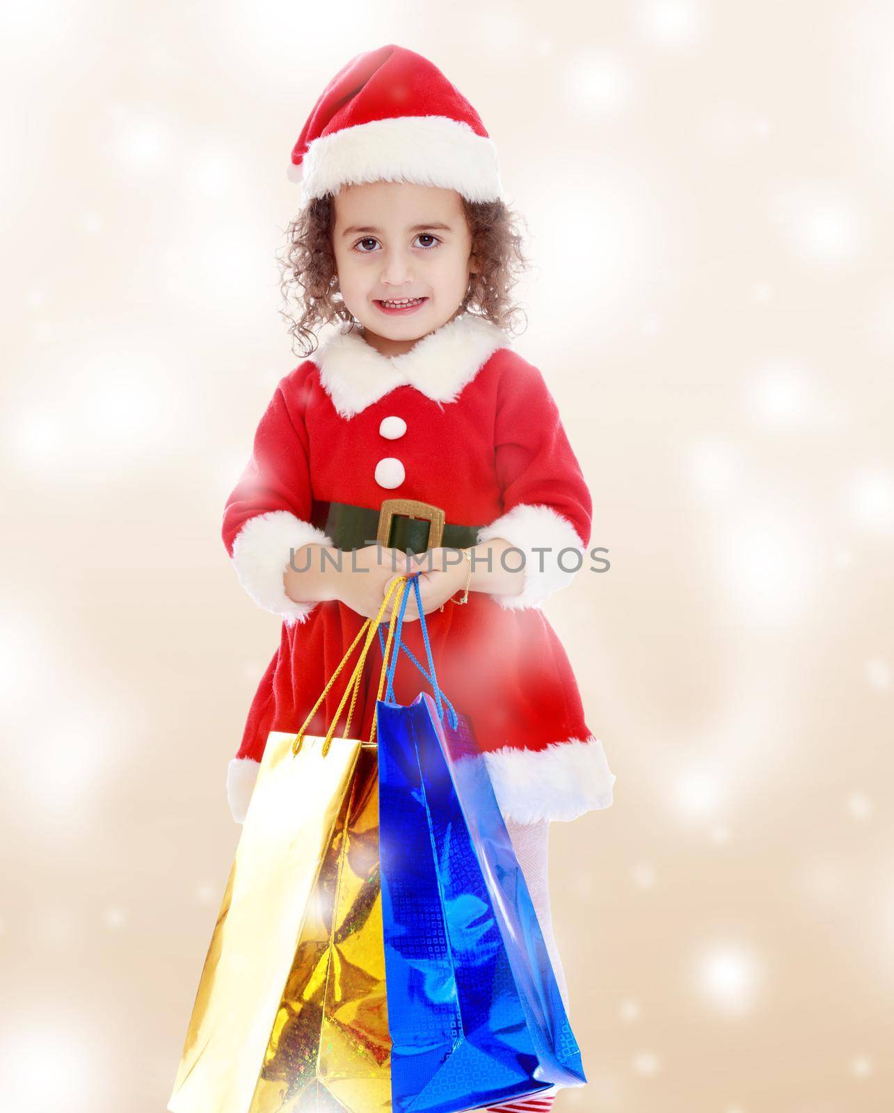 Gentle little curly-haired girl in a coat and hat of Santa Claus holding colorful shopping bags. Close-up.Winter brown abstract background with white snowflakes.