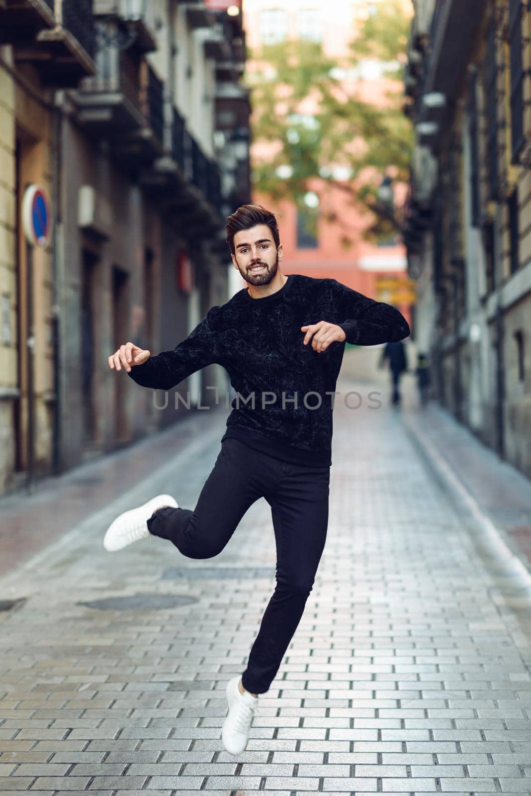 Young bearded man jumping in urban background with open arms wearing casual clothes. Guy with beard and modern hairstyle in the street.