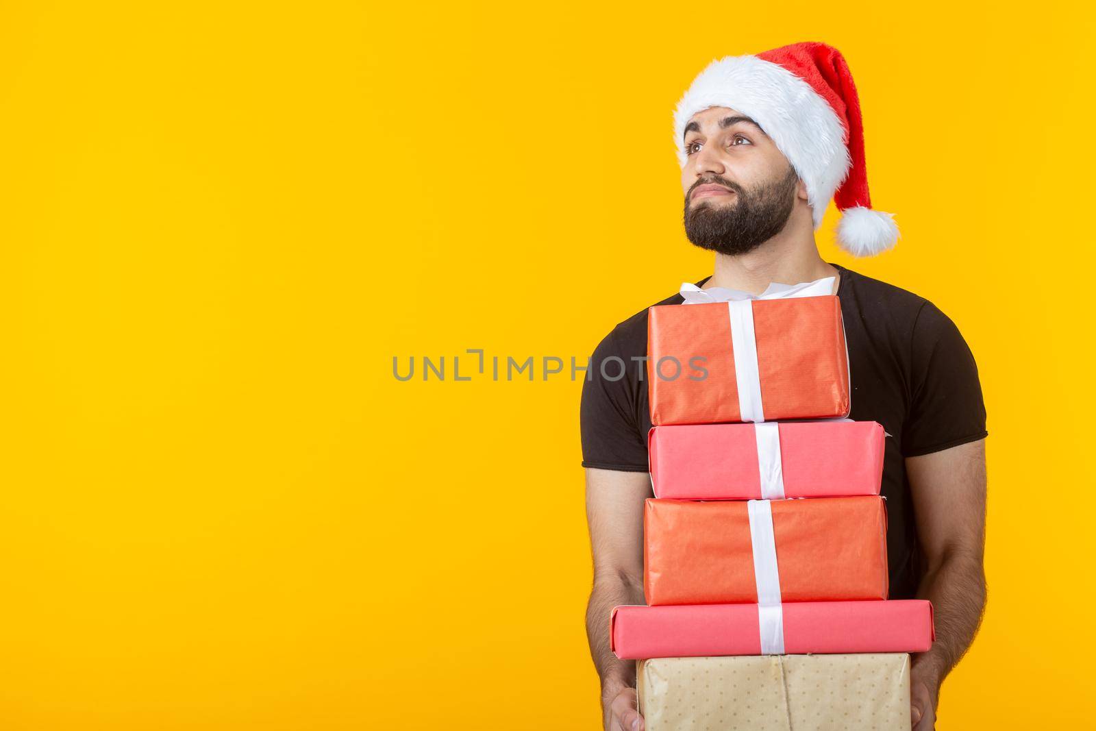 Disgruntled young man with a beard in a Santa Claus hat holds five gift boxes posing on a yellow background. Concept of gifts and greetings for Christmas and New Year