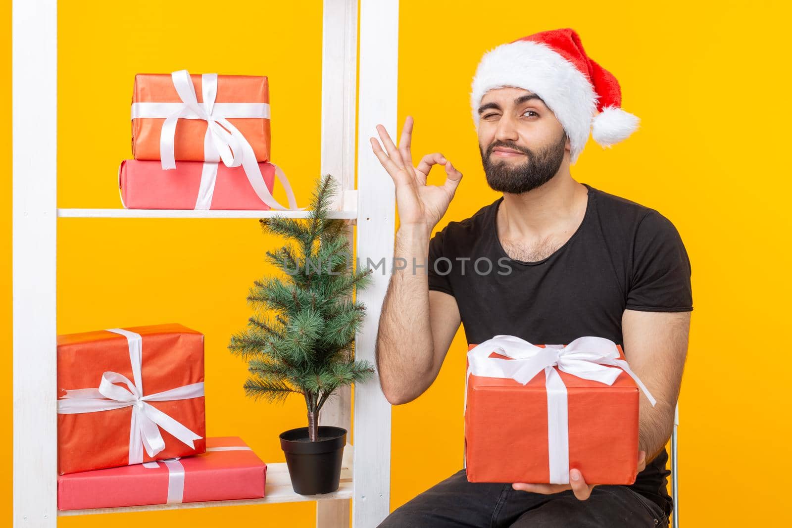 Young handsome man in a Santa Claus hat is holding congratulatory gifts next to a Christmas tree. Concept of holidays of christmas and new year