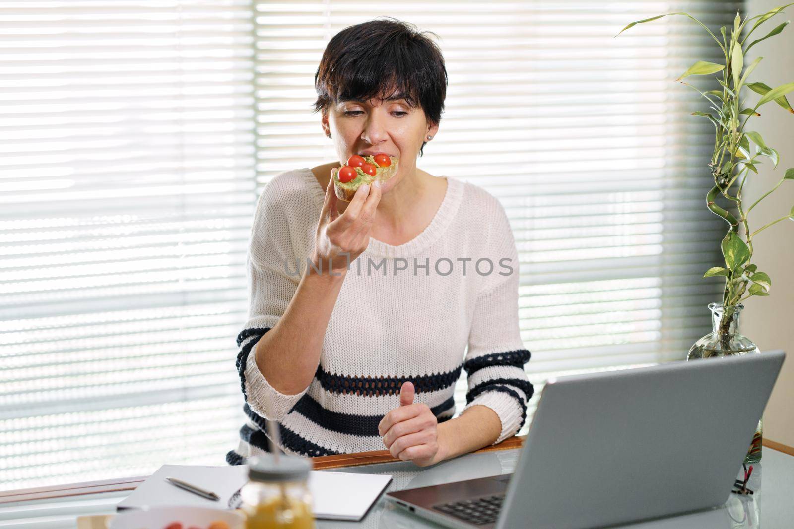 Woman eating some healthy food, while teleworking from home on her laptop. Female in her 50s