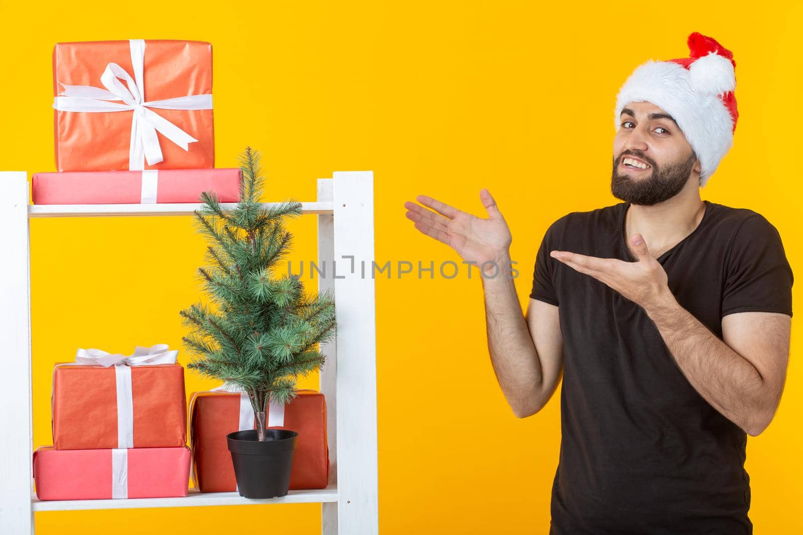 Young positive man with a beard and Santa Claus hat is standing near booth with gifts and Christmas tree on yellow background. Concept of congratulation gift and discounts for Christmas and New Year