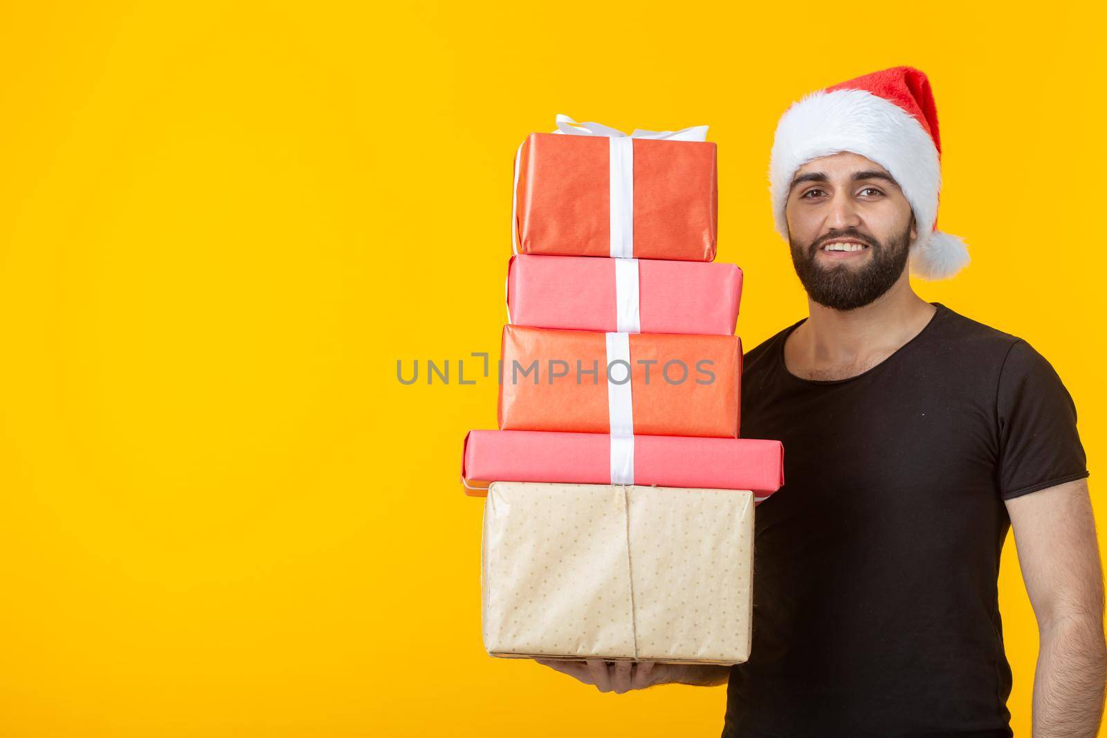 Disgruntled young man with a beard in a Santa Claus hat holds five gift boxes posing on a yellow background. Concept of gifts and greetings for Christmas and New Year