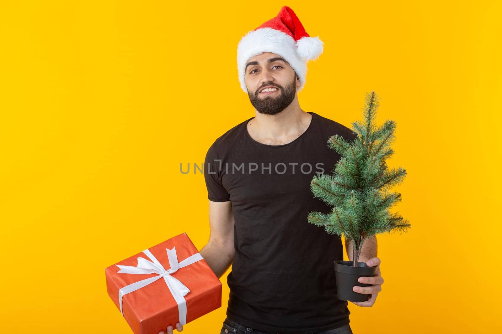 Handsome young man holding a red gift box and christmas tree in his hands posing in a New Year's cap on a yellow background. Merry Christmas and Happy New Year greetings concept