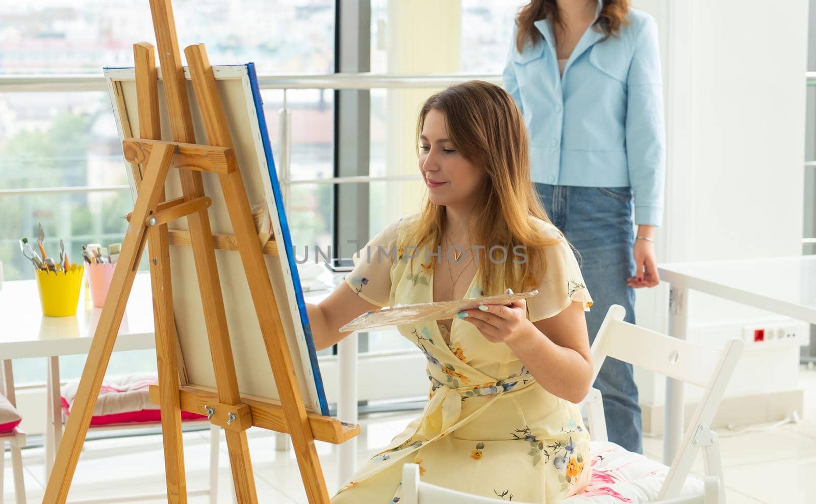 Art school, creativity and leisure concept - student girl or young woman artist with easel, palette and paint brush painting picture at studio. Teacher helps her by Satura86